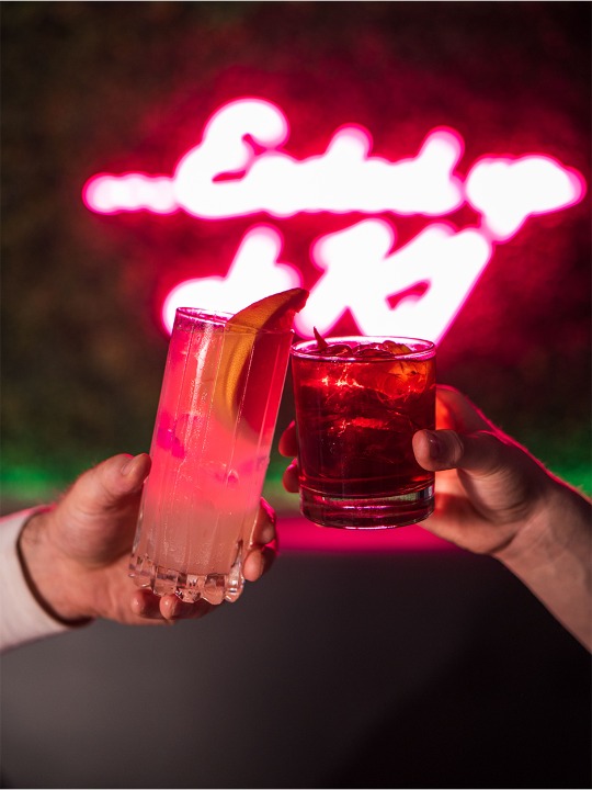 two people clink glasses with cocktails in front of a neon sign that reads" ended up at k1"